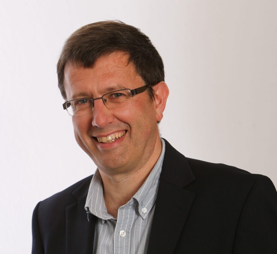 Head and shoulders photo of Alistair. He has short brown hair, and wears glasses and he is smiling widely