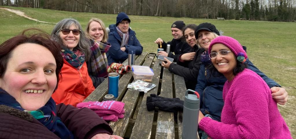 CCVS team get-together in spring 2023. Elli, Jigna, Lorna, Sally, Mark, and Chris are sitting at a wooden table in the countryside, wearing jumpers and hats. They are smiling and chatting.