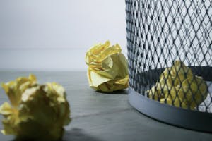 A paper bin with crumpled pieces of paper inside and some crumpled paper scattered outside of it.