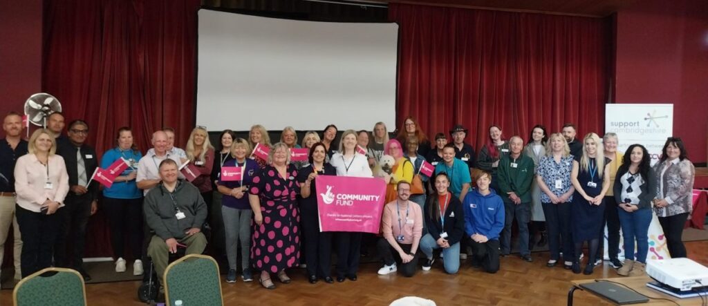 A diverse group of people stands together in a hall, smiling and holding a pink "Community Fund" banner, at the launch of the Support Fenland project