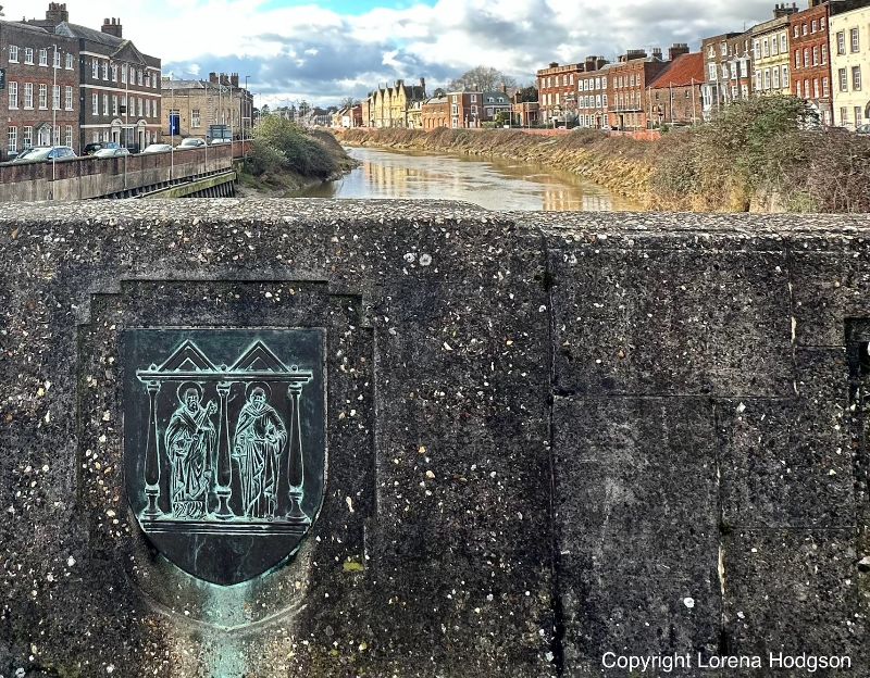 Photo of A view of the famous North and South Brink from the Town Bridge, with the town crest. Riverside buildings line the river Nene including Octavia Hill Birthplace House, Sessions House, and Peckover House.