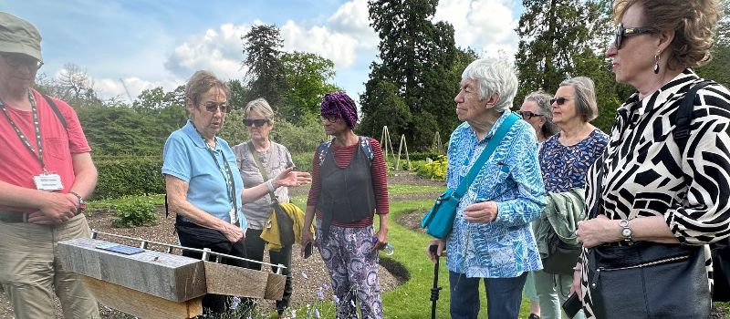 A group gather together to talk about the flowers in a beautiful flower bed in a lush and rich garden. The sky is bright blue and the group are enjoying their visit and have smiles on their faces. 