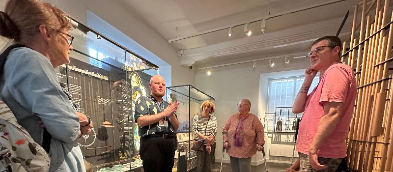 A group of people stand and listen to a speaker inside a museum filled with glass cabinets protecting pictures and pottery artefacts which have been collected throughout Ely. 