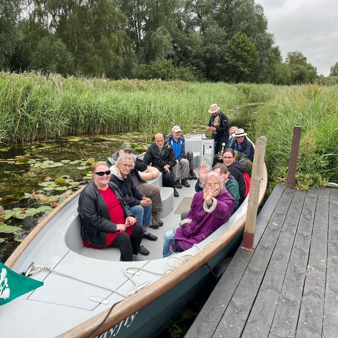 A group of people are sitting in a boat moored at a boarding pontoon, they are waiting along the riverbank which is filled with high reeds. The water is green in colour and the individuals in the boat are wearing coats, and one lady has dark glasses on.