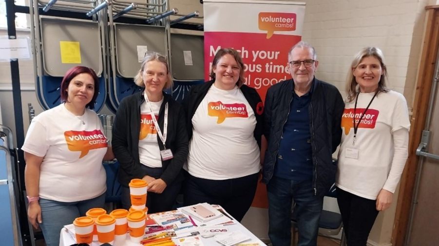 Five people stand smiling behind a table with brochures and branded items. A "Volunteer Cambs" banner is in the background