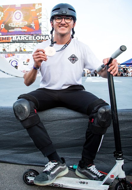 Jaden Sharman sitting on the edge of a large padded mat, he wears a white tshirt and back trousers with knee safety pads. He holds a scooter in one hand, and a silver medal in the other, which is on a ribbon round his neck.