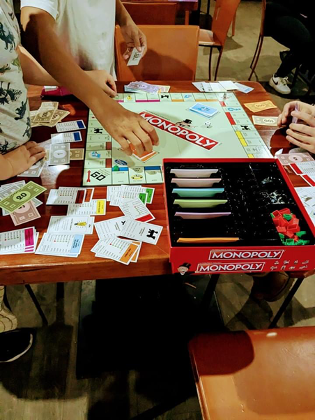 A monopoly board game in progress. Hands hold the game money and properties, and one young person has just picked up a game card