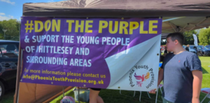 A young man standing beside a purple banner that says - Don The Purple and support the young people of Whittlesey and surrounding areas.