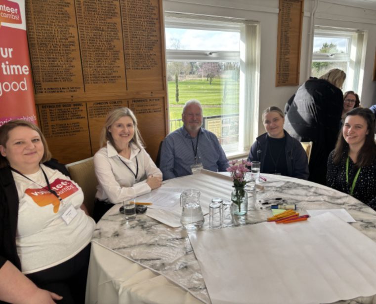 A group of people sitting round a table at the breakfast lunch smiling, there is a jug of water and glasses on the table and coloured pens and paper
