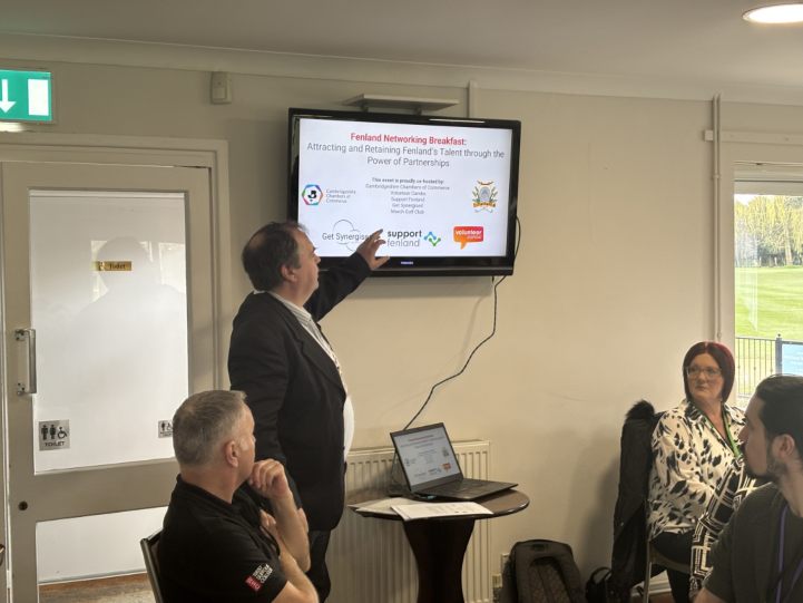 A man giving a presentation using a wall mounted screen to show slides at the networking breakfast while the attendees listen 