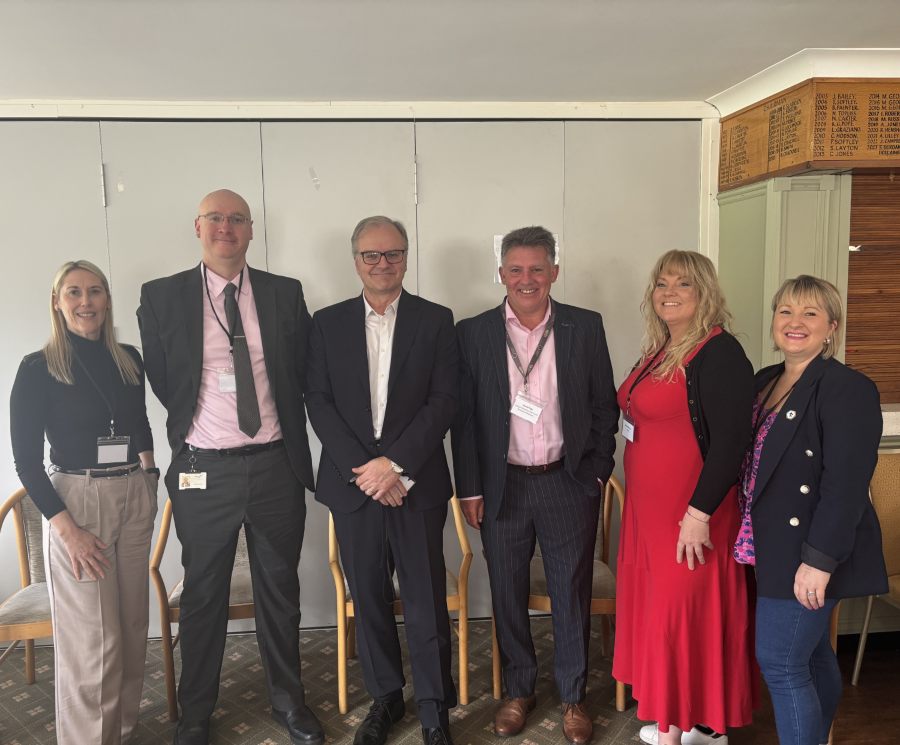 A group of six people, three men and three women, stand smiling in a row at the networking breakfast. The tone is professional and friendly, conveying teamwork. 