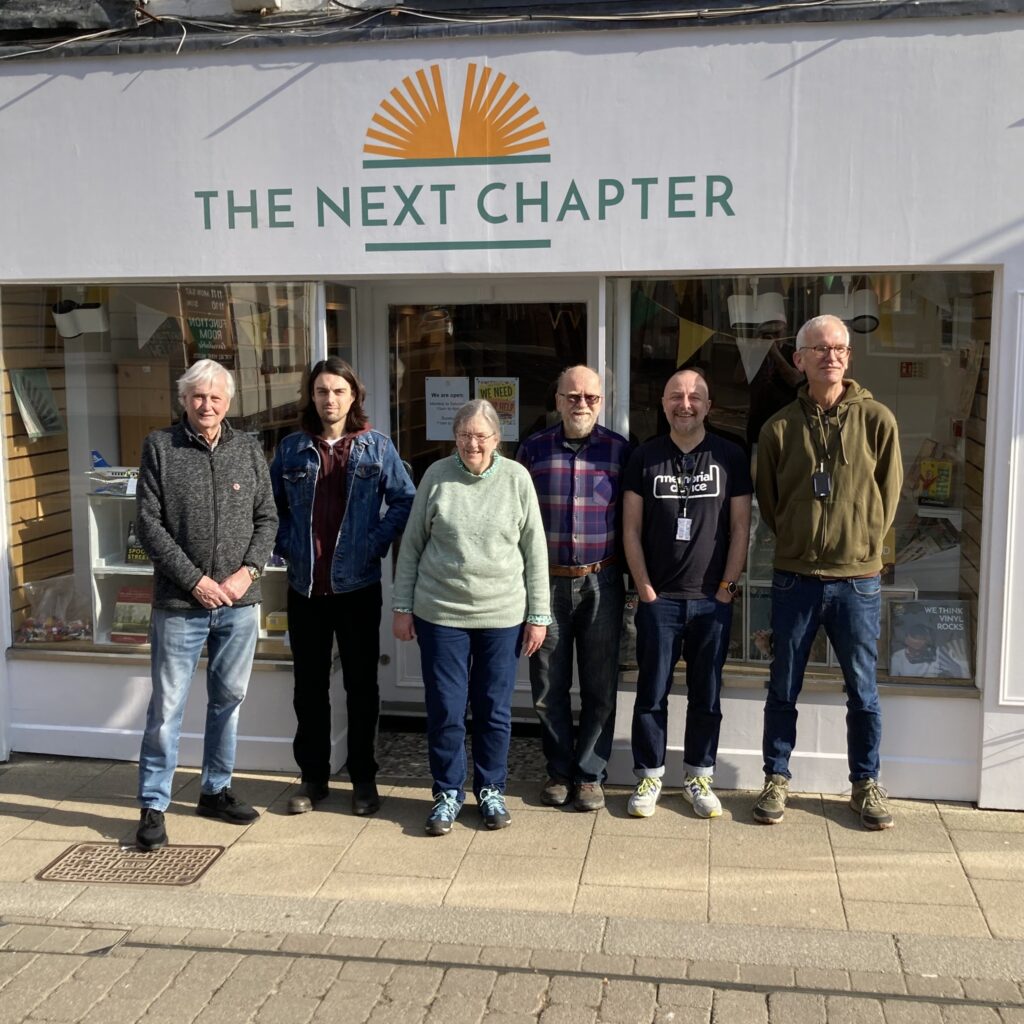Six people standing outside a shop called "The Next Chapter," posing for a group photo on a sunny day.