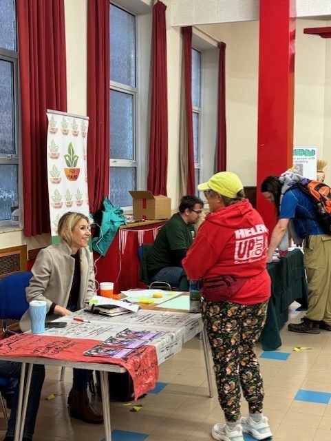 A woman in a red hoodie talks to a seated woman at an indoor event. There is a bright, friendly atmosphere with colourful banners.