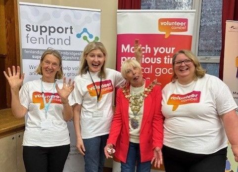 Four women stand smiling in front of banners for volunteer organisations. Three wear "Volunteer Cambs" shirts