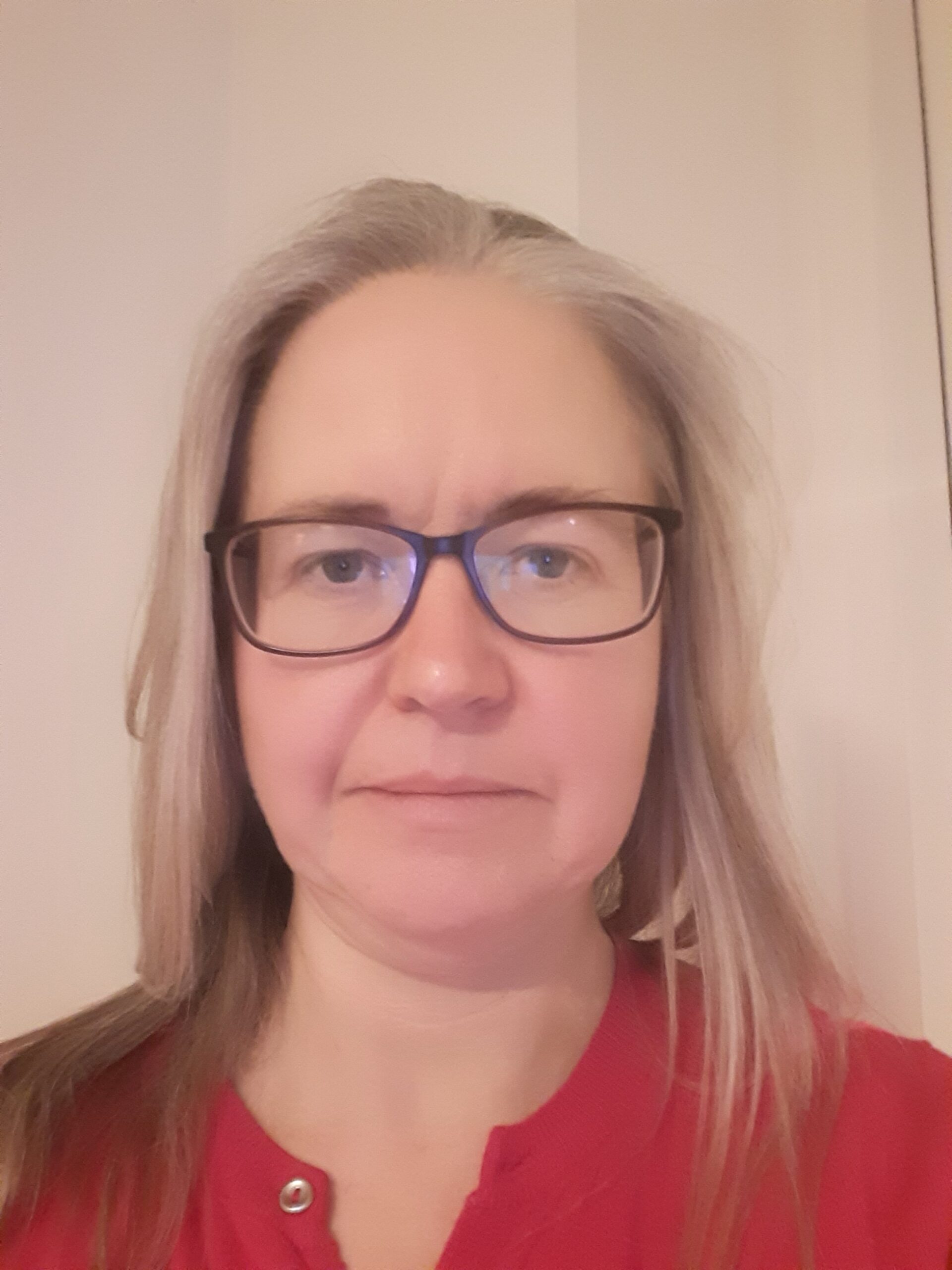 A head and shoulders photo of Anne. Ann has blond shoulder length hair and wears glasses. She is wearing a red top