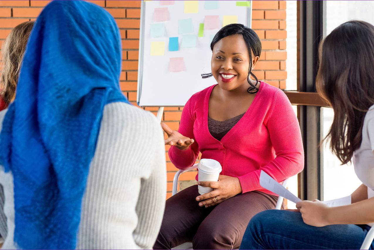 A group of 4 women sit in a circle talking, telling each other stories. A black woman sits facing the camera, she is wearing a bright pink jumper and brown trousers, she is smiling widely as she holds her had towards the group as she talks.