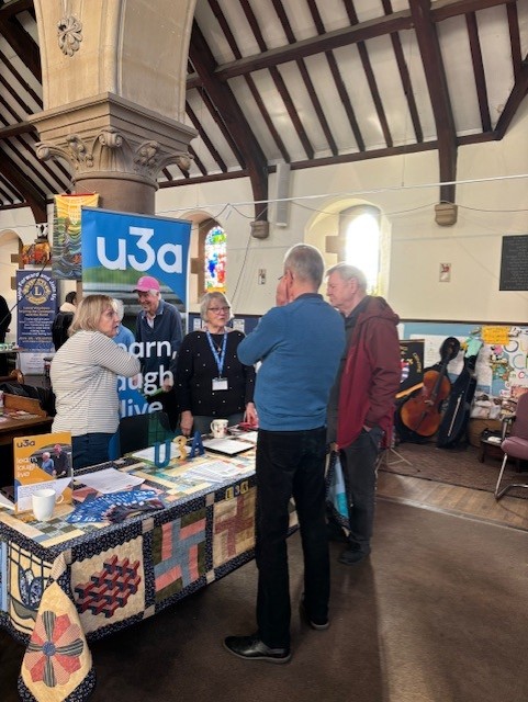 Two people stand and chat to stallholders at The Volunteer fair. The stall is covered with leaflets and information.