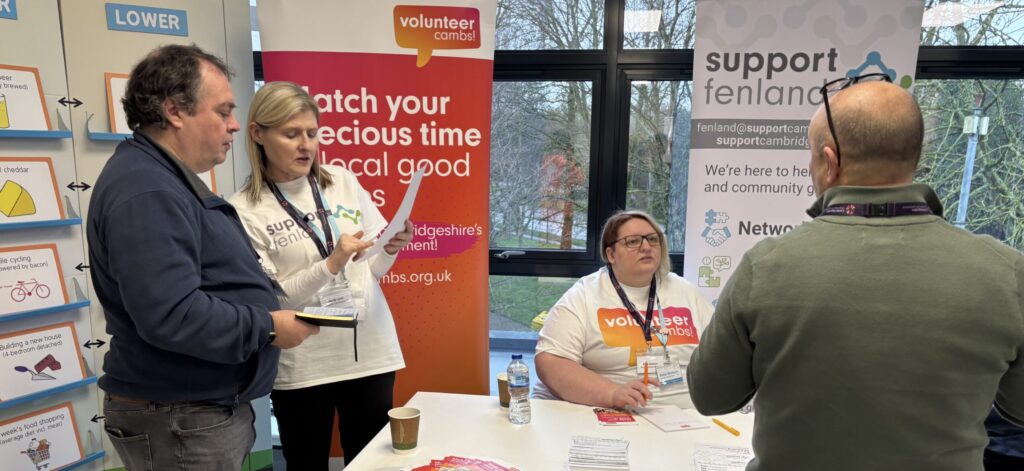 Two people standing and speaking with staff at a Support Fenland and Volunteer Cambs information table, which is set up with leaflets and banners in the background.