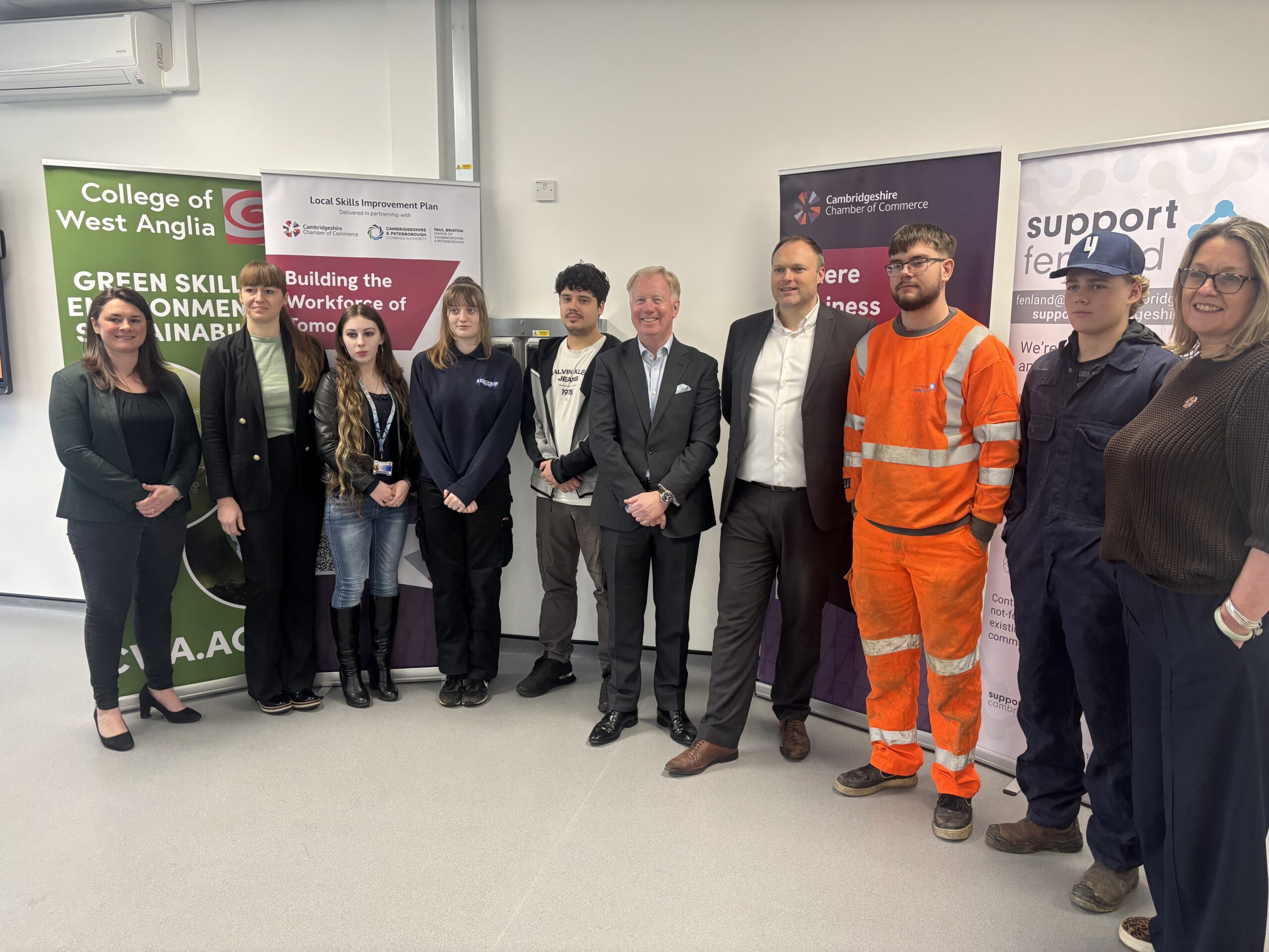 A group of people standing indoors in front of organisational banners, including the College of West Anglia and Support Fenland. Some are in work uniforms and others in business or casual clothing.