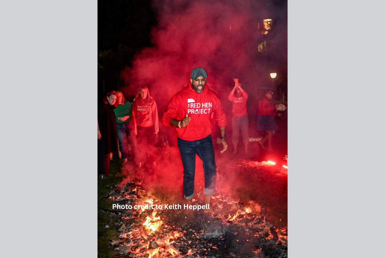 A man runs across hot coals at a fundraising firewalk event, he has in mouth open in a shout. There are falmes and smoke coming from the coals